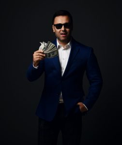 Portrait of insolent rich businessman in blue jacket, white shirt and sunglasses standing holding bundle, fan of dollars cash and the other hand in pocket of his pants over dark background